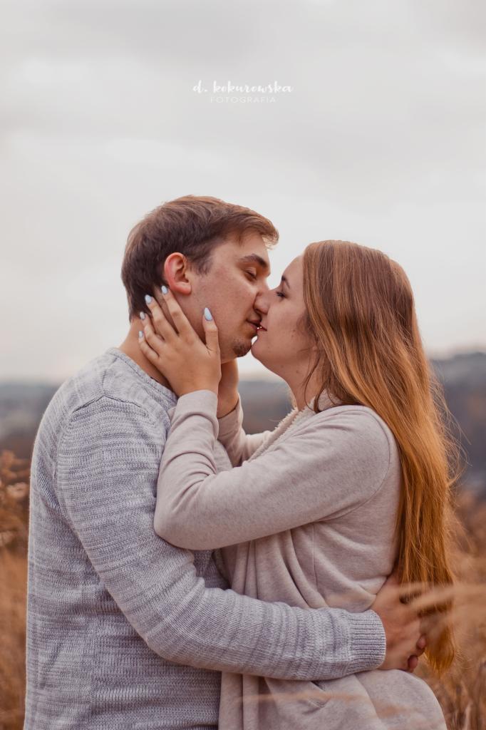 Zakochana para całuje się na tle jesiennego krajobrazu; kobieta z długimi, rudymi włosami dotyka twarzy mężczyzny z krótkimi, brązowymi włosami, oboje ubrani w swetry w odcieniach szarości.