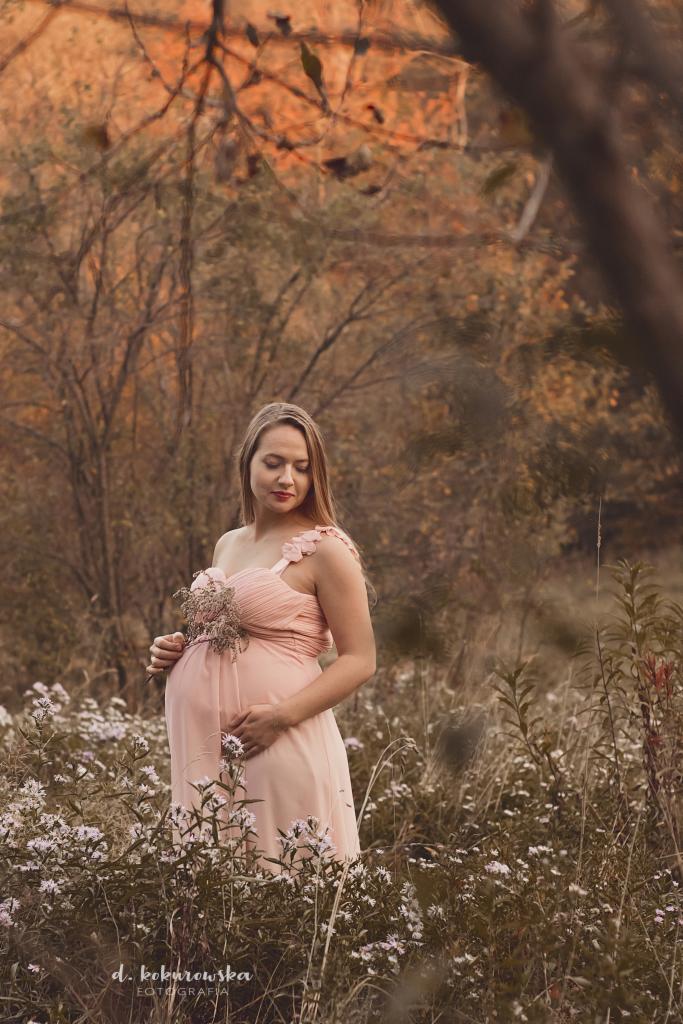 Kobieta w ciąży w zwiewnej, pudrowo-różowej sukience, stojąca na łące z białymi kwiatami, trzymająca bukiecik suszonych kwiatów, sfotografowana w jesiennej scenerii z drzewami w tle.
