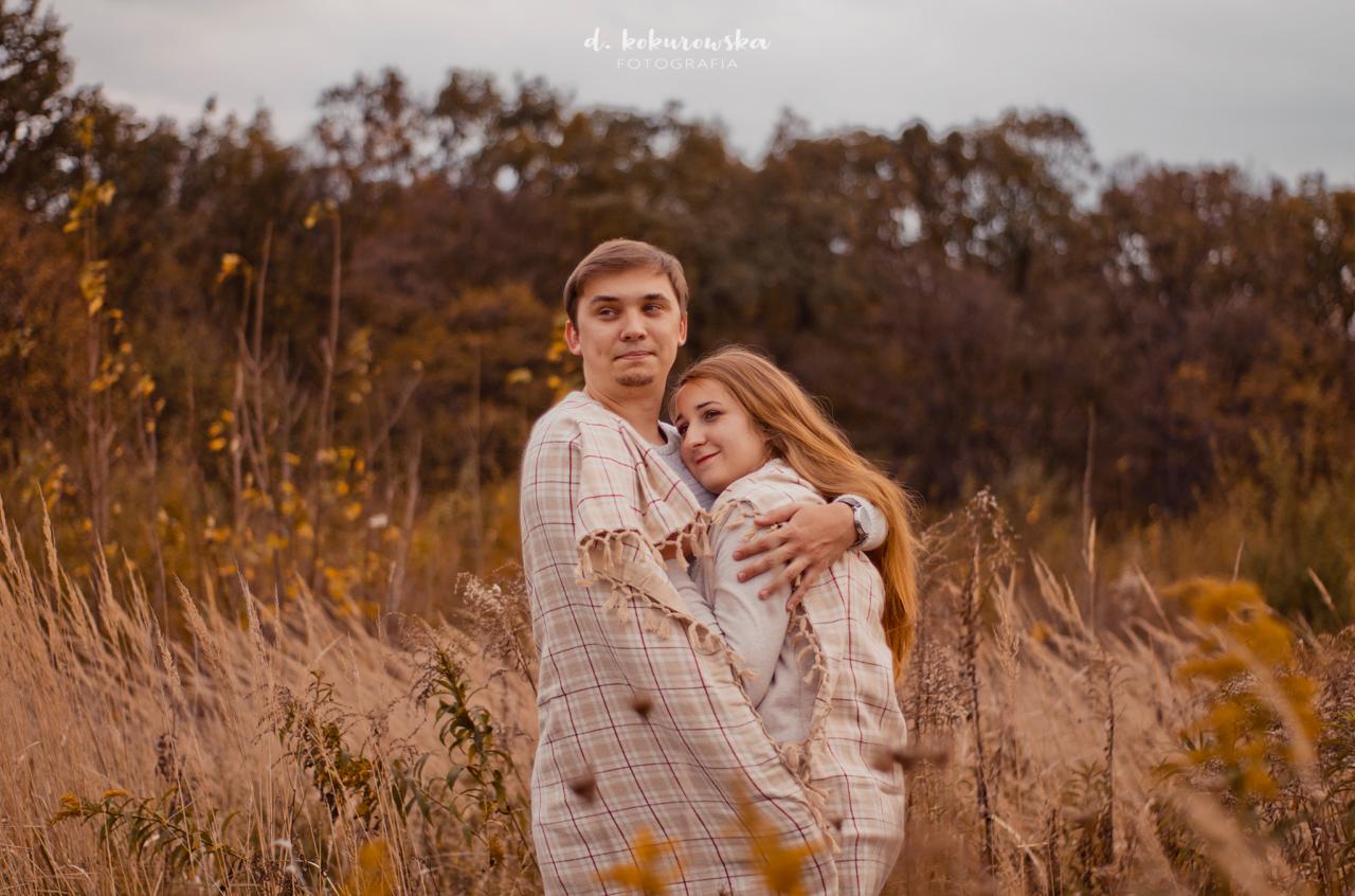 Para otulona pledem w kratę, obejmująca się na tle jesiennego krajobrazu z wysoką trawą i drzewami, w tle widoczne logo fotografa.