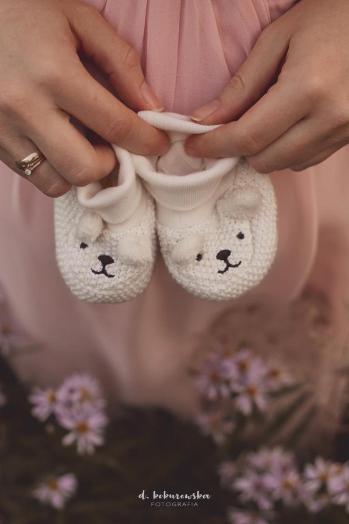Dłonie kobiety z pierścionkiem trzymają białe, dziergane buciki niemowlęce z motywem niedźwiedzia na tle pudrowo-różowej sukienki i rozmytych kwiatów.