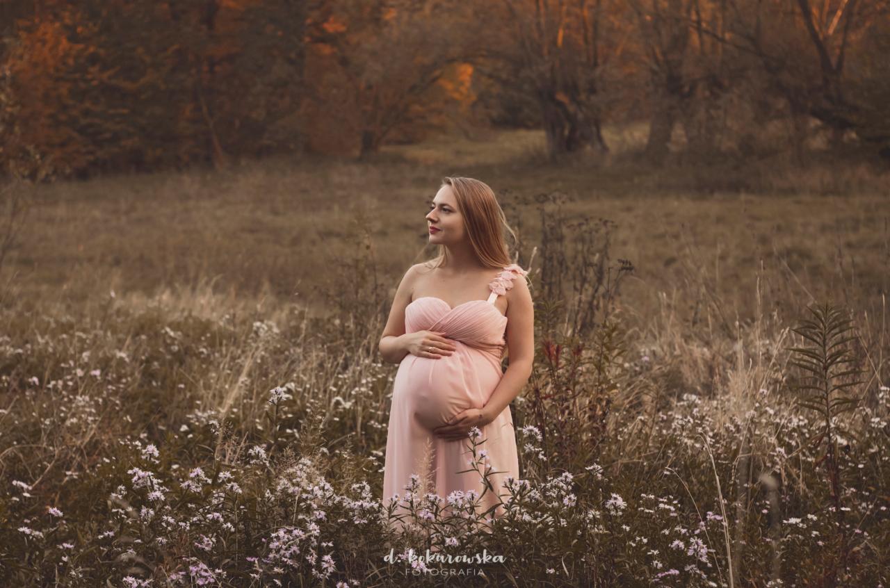 Kobieta w jasnoróżowej sukni ciążowej stoi na łące pełnej białych kwiatów, z zamyślonym wyrazem twarzy spogląda w dal, jesienne drzewa w tle.