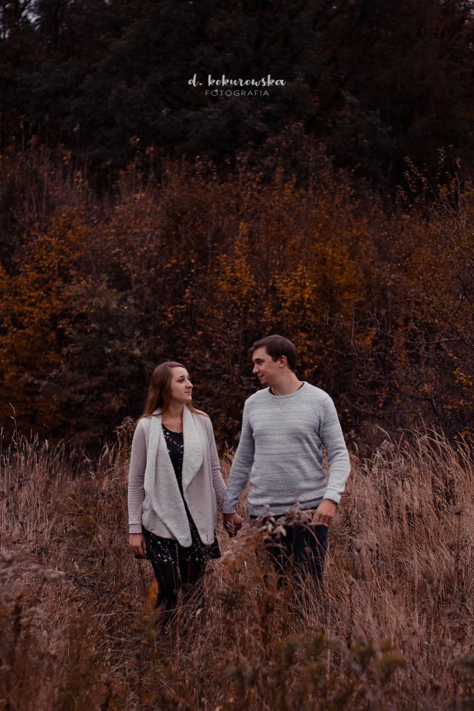 Para trzymająca się za ręce spaceruje po polu z wysoką trawą w jesiennej scenerii, delikatne spojrzenia, logo fotografa w górnej części zdjęcia.