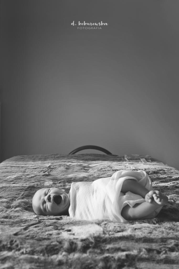 Czarno-białe zdjęcie noworodka owiniętego w białą tkaninę, leżącego na teksturowanym kocu z otwartymi ustami, sugerujące ziewanie; w tle widoczna rama łóżka i logo fotografa.