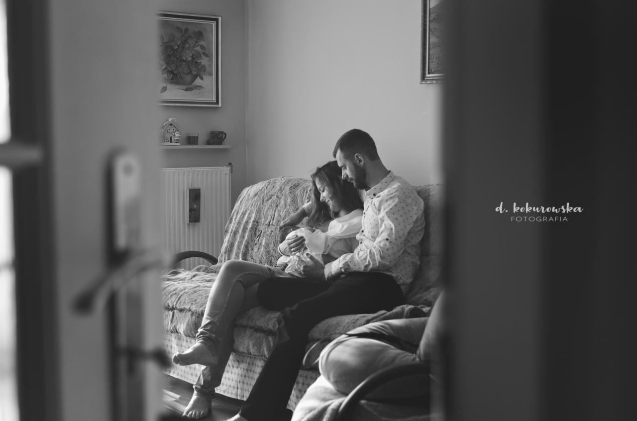 Czarno-białe zdjęcie przedstawia parę siedzącą na kanapie w pokoju, trzymającą noworodka. Kobieta w jasnej bluzce i dżinsach, mężczyzna w koszuli w drobne wzory i ciemnych spodniach. W tle widoczne...