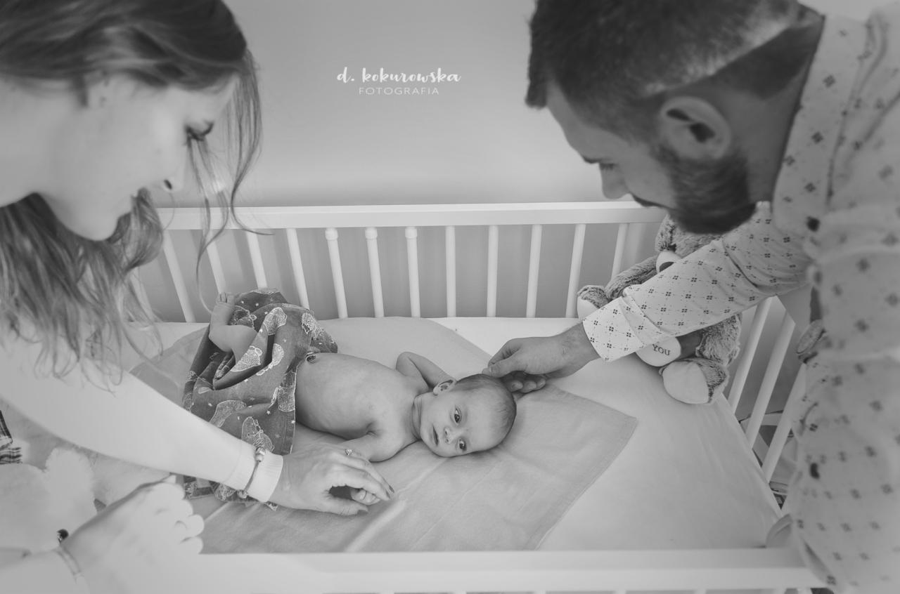 Czarno-białe zdjęcie z Cieszyna przedstawia rodziców pochylających się nad noworodkiem leżącym w białym łóżeczku, obok pluszowy miś.