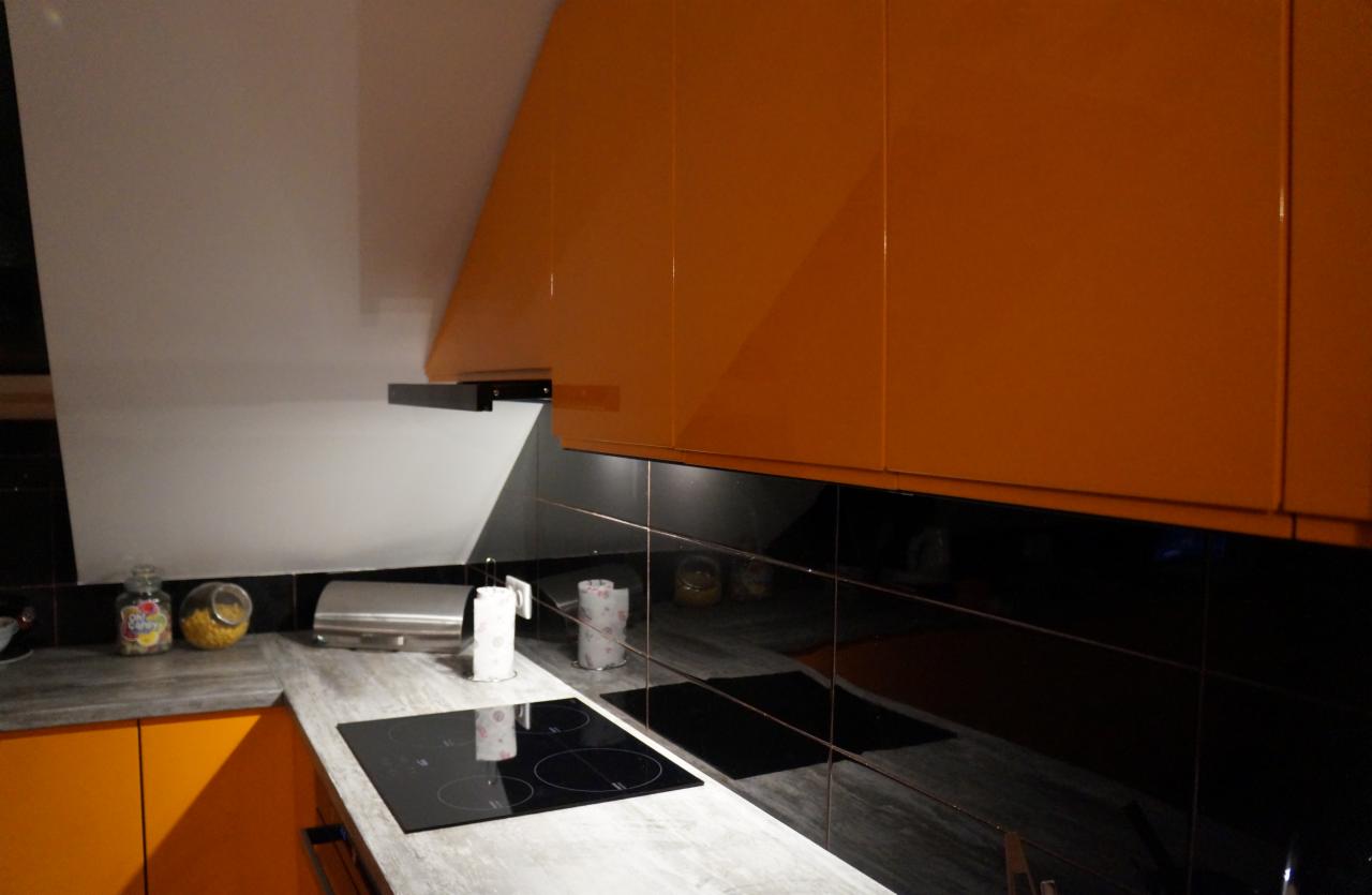 Pomarańczowe, lakierowane szafki kuchenne wiszące nad czarnymi, błyszczącymi płytkami, widoczny fragment blatu z płytą indukcyjną i akcesoriami.