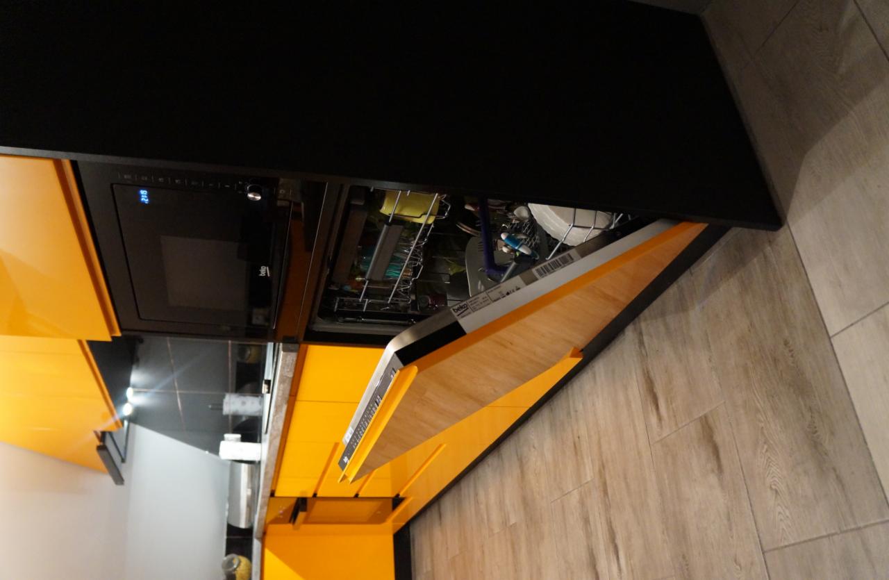 Żółta zabudowa kuchenna z otwartą zmywarką, widoczne naczynia wewnątrz, czarny piekarnik w zabudowie powyżej, drewnopodobna podłoga.