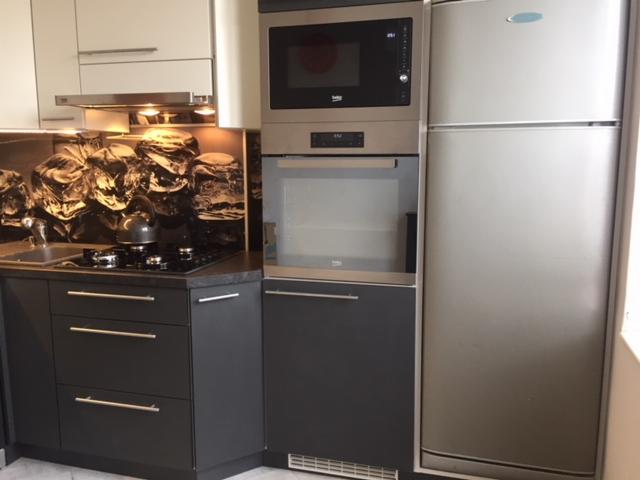 Część zabudowy kuchennej z szarymi szafkami z trzema szufladami, piekarnikiem i mikrofalówką w zabudowie oraz srebrną lodówką; na blacie czajnik i płyta gazowa, a na ścianie panel dekoracyjny...