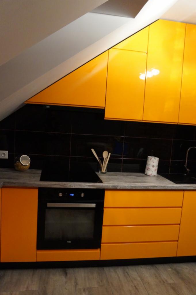 Żółte szafki kuchenne pod skosem dachu, czarny blat i płytki, widoczny piekarnik i akcesoria kuchenne.