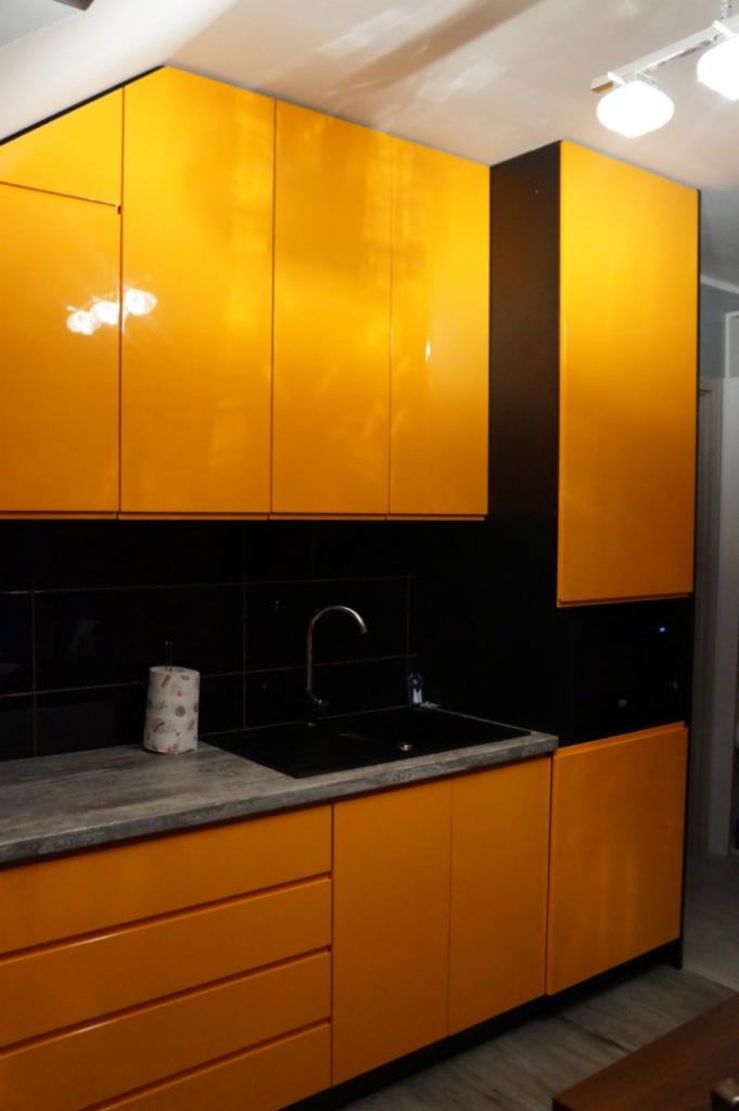 Żółta, lakierowana zabudowa kuchenna z czarnym blatem i płytkami, widoczny zlew i bateria.