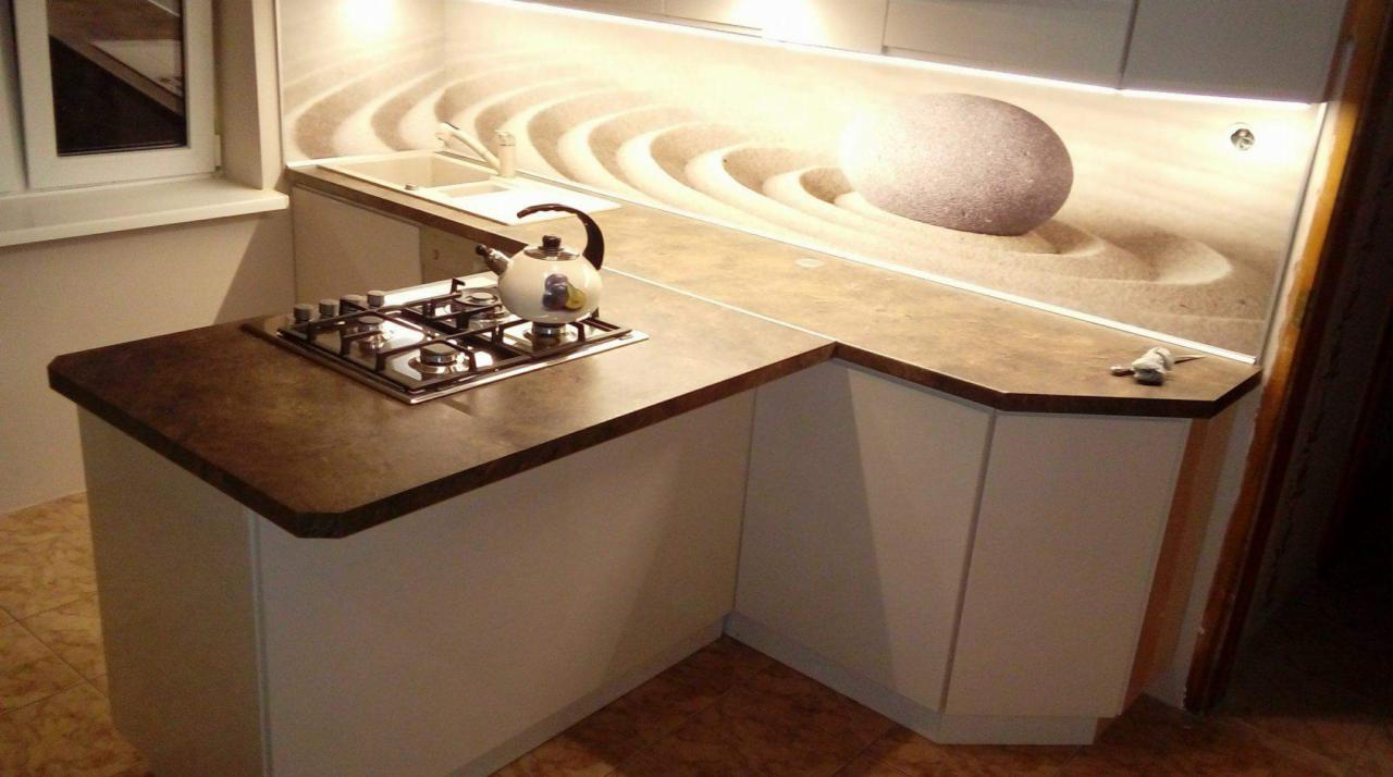 Kuchenny blat z brązowego konglomeratu, płyta gazowa z czajnikiem i panel szklany z motywem zen, oświetlenie LED pod szafkami.