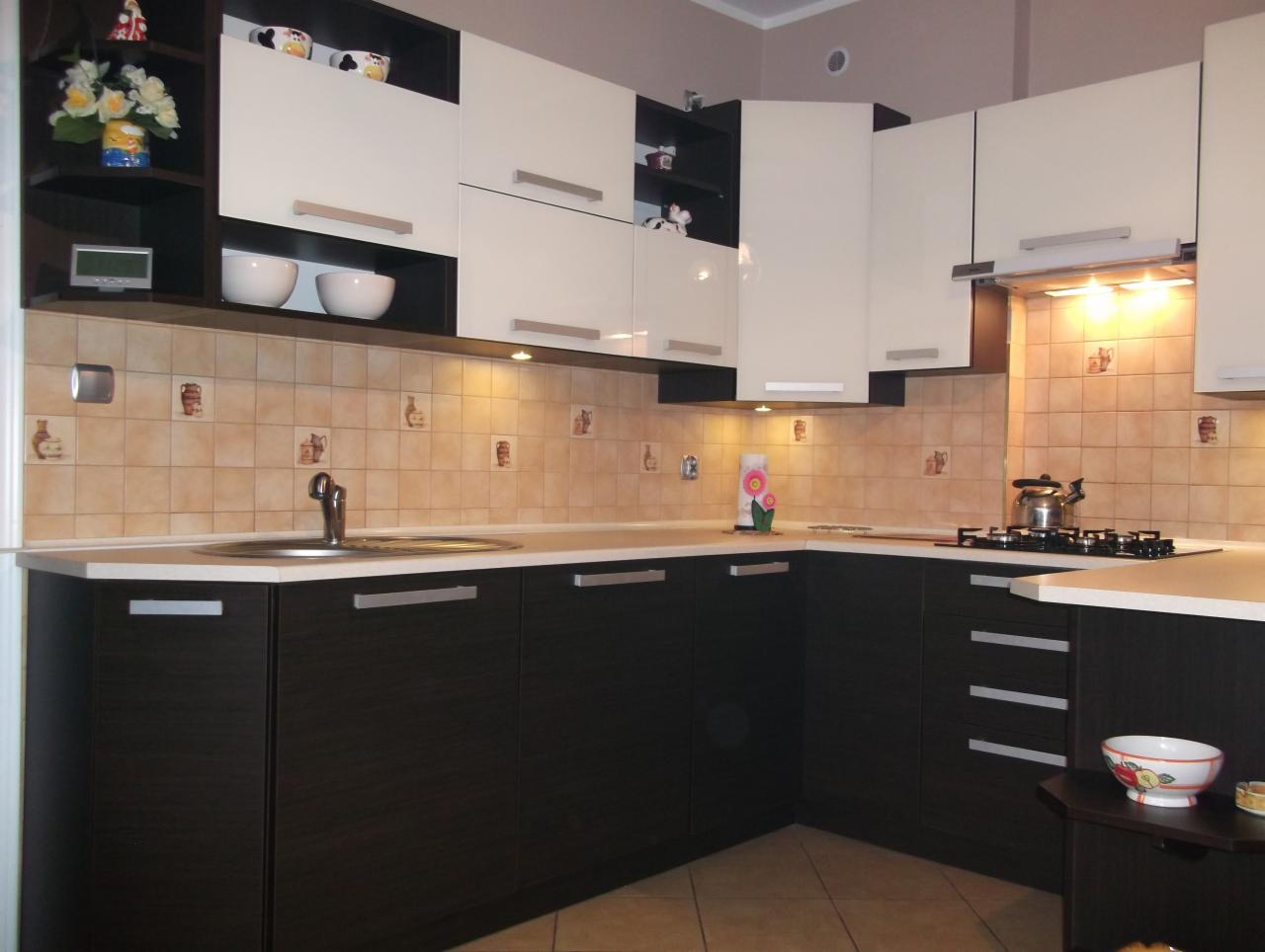 Dwukolorowa zabudowa kuchenna z ciemnobrązowymi dolnymi szafkami i białymi górnymi szafkami, widoczne płytki ceramiczne z motywem rustykalnym i podświetleniem podszafkowym.