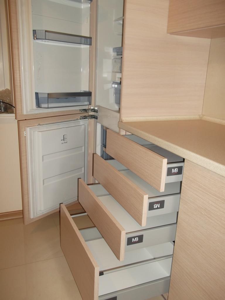 Jasna szafa na wymiar z trzema wysuwanymi szufladami z frontami z jasnego drewna, zintegrowana z lodówką, widoczne logo 'MB' na szufladach.