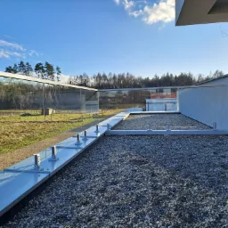 Szklana balustrada z metalowymi mocowaniami na dachu pokrytym żwirem, w tle widok na las i błękitne niebo z chmurami.