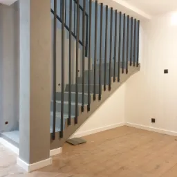 Schody betonowe z czarną, minimalistyczną balustradą z pionowych prętów, widok na drewnianą podłogę i białe ściany z czarnymi gniazdkami.