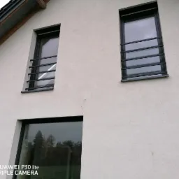 Elewacja domu z oknami zabezpieczonymi poziomymi, czarnymi kratami. Widok z dołu, ukazujący dwa okna na poddaszu i fragment okna na parterze. Tynk zewnętrzny w odcieniu jasnej szarości.