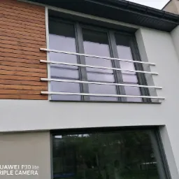 Nowoczesna fasada budynku z drewnianą okładziną i białym tynkiem, z oknem zabezpieczonym poziomą, metalową konstrukcją przypominającą balustradę.