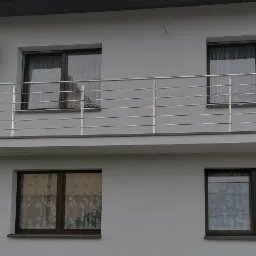 Szczegółowe ujęcie balkonu z prostą, nierdzewną balustradą w Orzeszu, zamontowaną na tle szarej elewacji budynku z widocznymi oknami i anteną satelitarną.