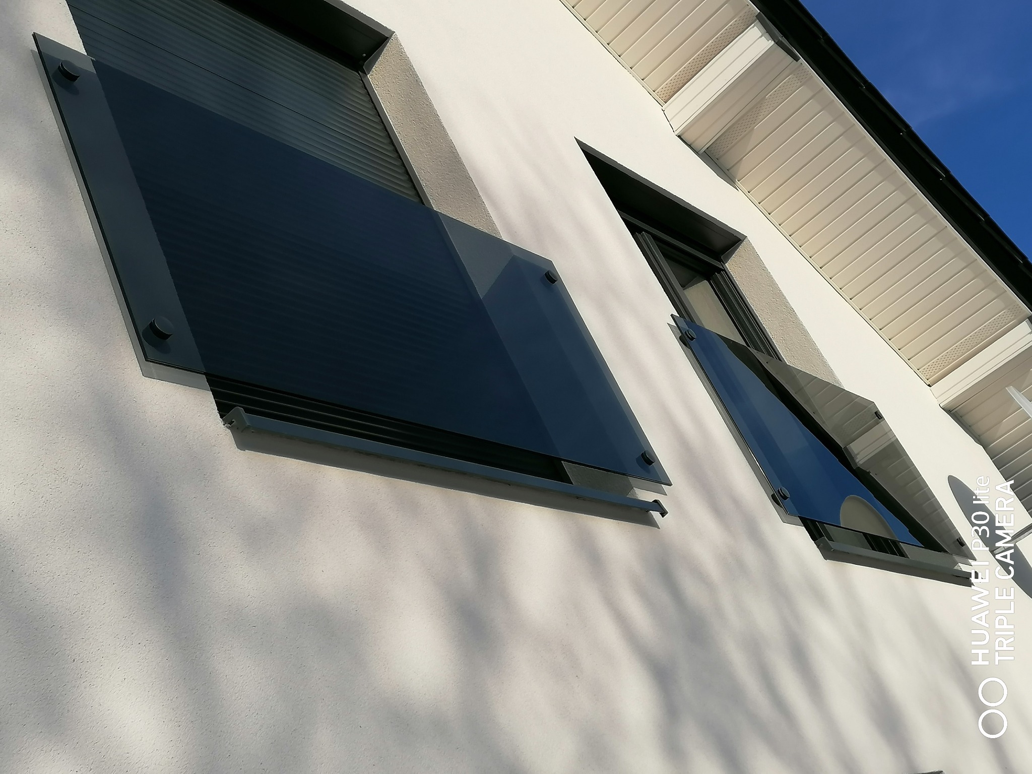 Nowoczesne, szklane balustrady okienne zamontowane na białej elewacji budynku, widoczne mocowania punktowe i refleksy nieba na szkle.