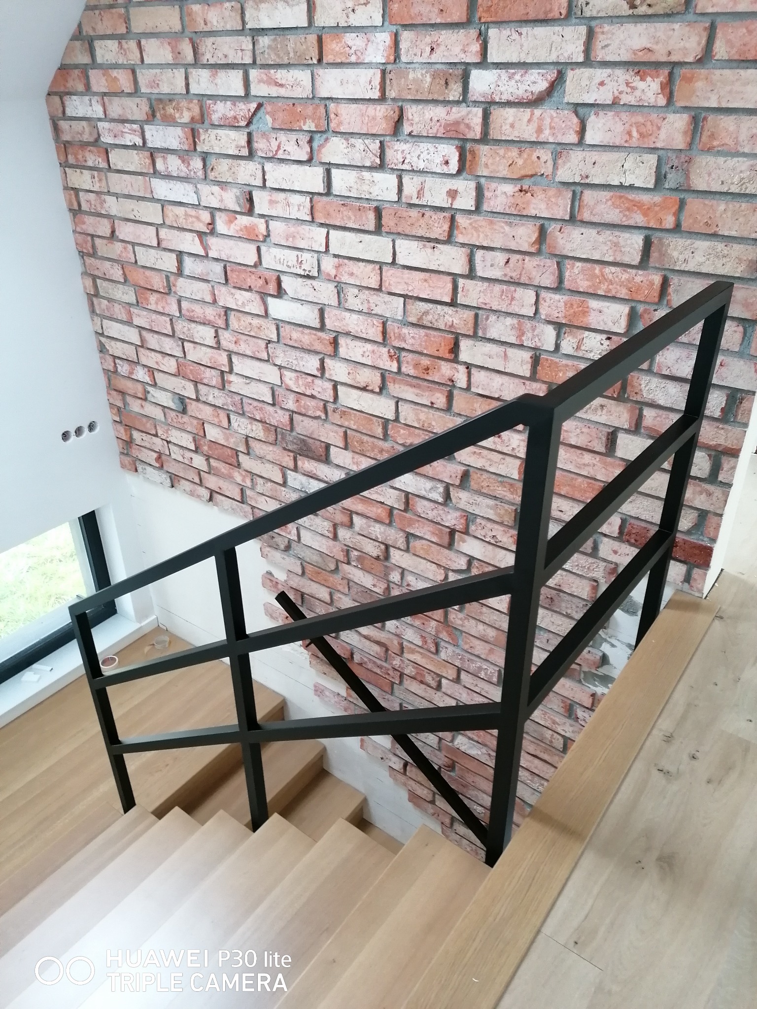 Wnętrze domu: schody drewniane z czarną, metalową poręczą na tle ściany z cegły dekoracyjnej, widok z góry.