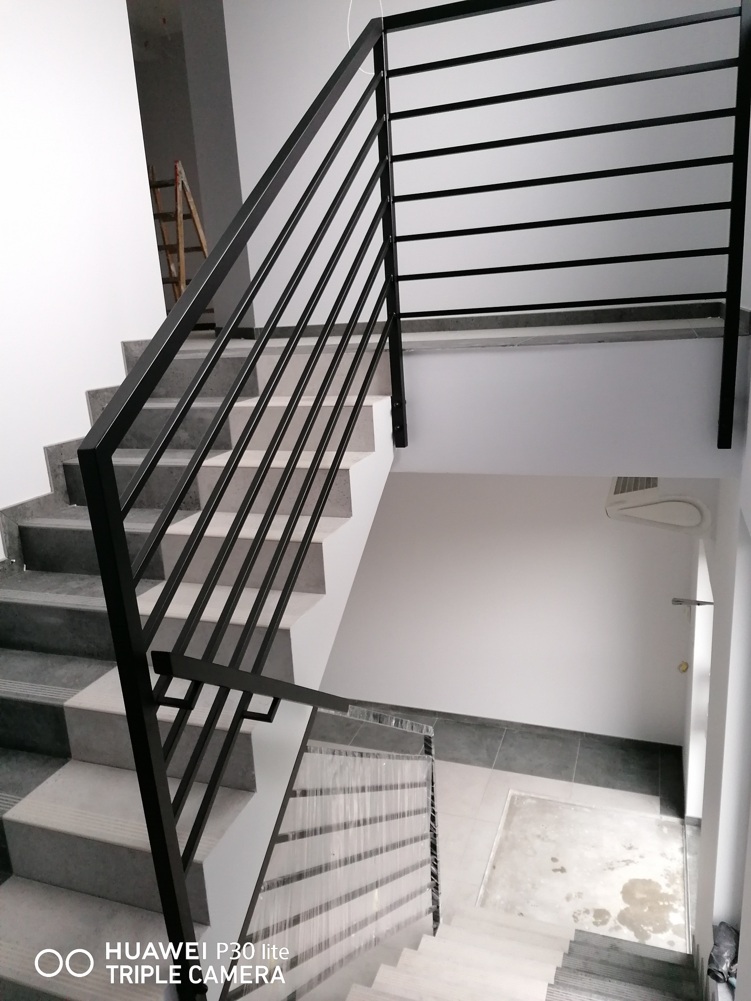 Wewnętrzne schody z szarymi stopniami wykończone czarną, metalową balustradą z poziomymi prętami, widok z góry na klatkę schodową w trakcie wykończenia.