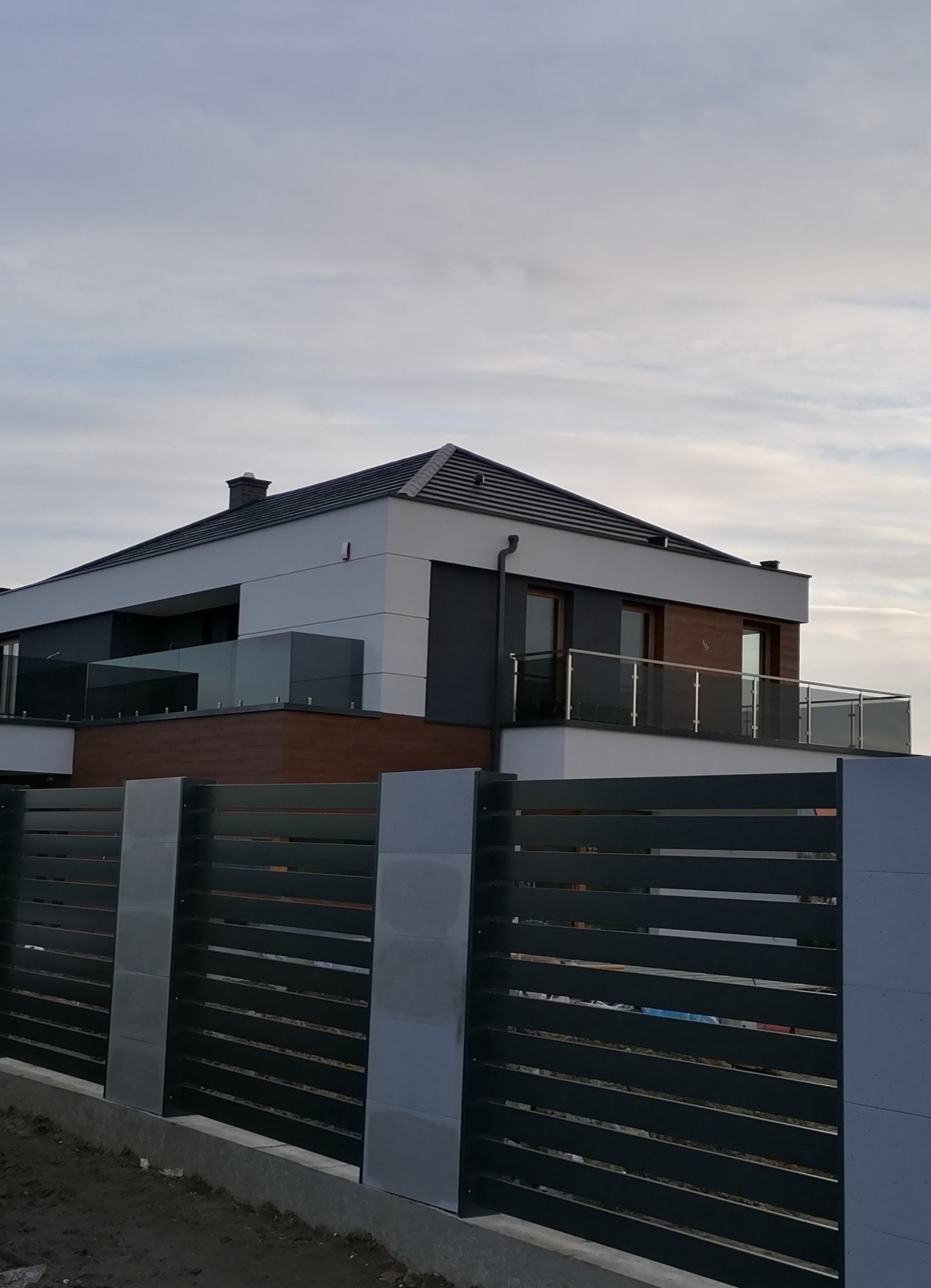 Nowoczesny dom z szarą, poziomą, lamelową bramą wjazdową i szklanymi balustradami balkonowymi, widoczne elementy wykończenia elewacji imitujące drewno.