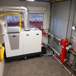 Nowoczesny biały piec grzewczy De Dietrich z żółtymi rurami gazowymi w pomieszczeniu z szarymi ścianami z blachy trapezowej i licznymi instalacjami hydraulicznymi.