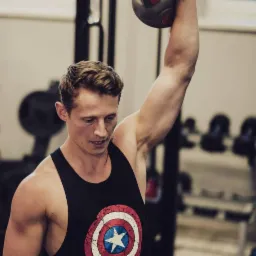 Młody mężczyzna w czarnym tank topie z motywem Kapitana Ameryki, unoszący piłkę lekarską nad głową w siłowni.