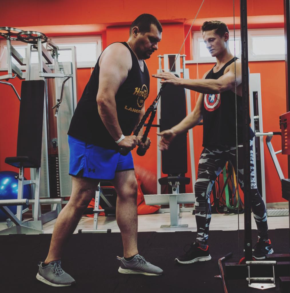 Mężczyzna w czarnej koszulce z logo Hard Rock Cafe ćwiczy na maszynie pod okiem instruktora w koszulce z motywem Kapitana Ameryki, obaj w sportowych strojach na tle siłowni z pomarańczowymi ścianami.