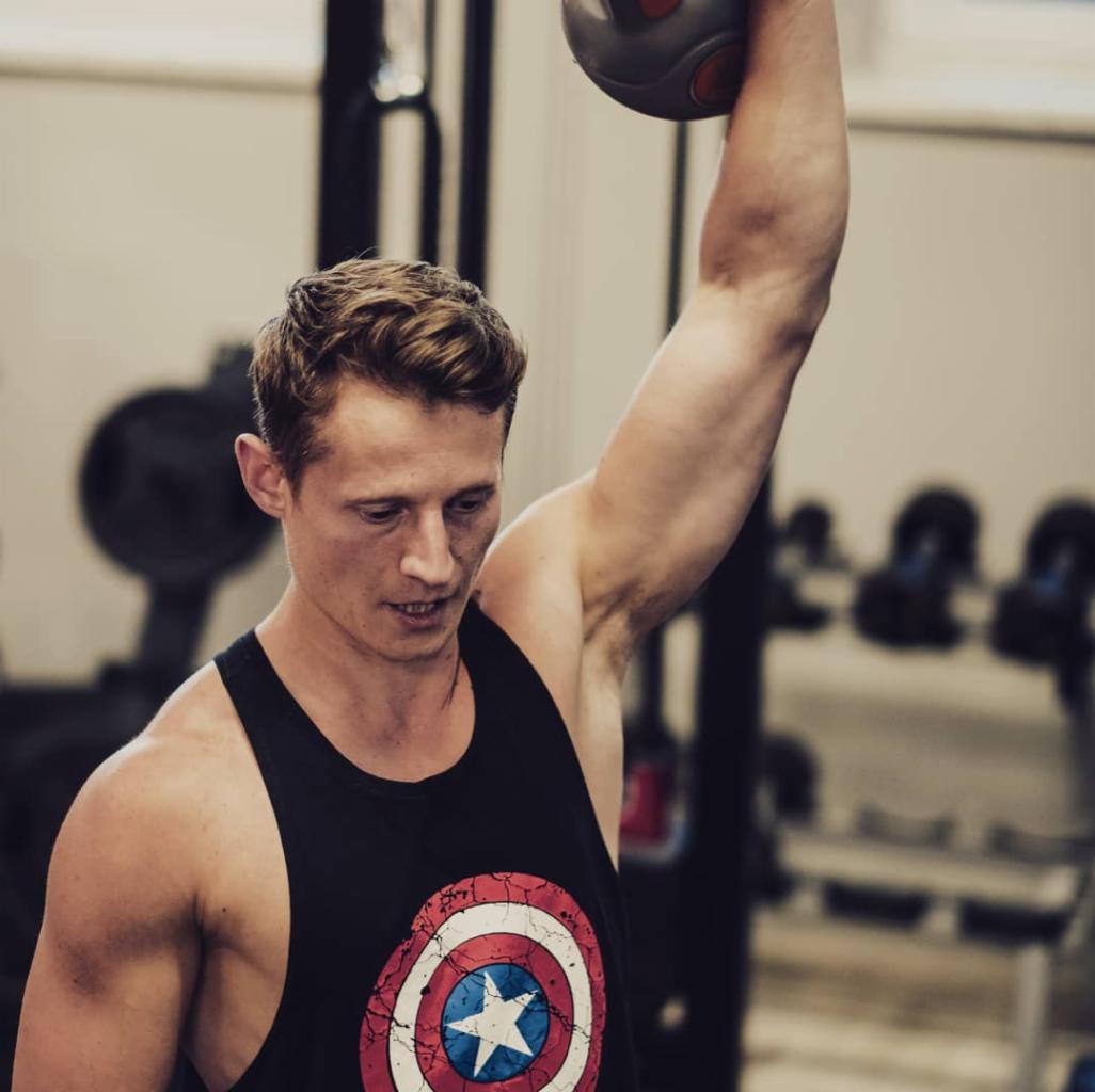 Młody mężczyzna w czarnym tank topie z motywem Kapitana Ameryki, unoszący piłkę lekarską nad głową w siłowni.