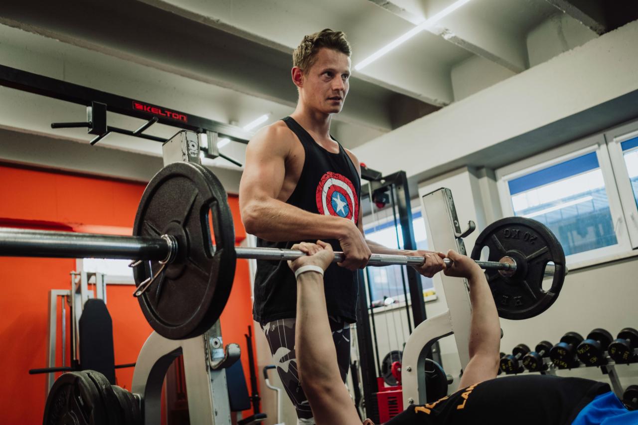 Mężczyzna w koszulce z motywem Kapitana Ameryki asekuruje ćwiczącego mężczyznę podczas wyciskania sztangi na ławeczce w siłowni.