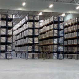 Wnętrze magazynu wysokiego składowania z regałami wypełnionymi zapakowanymi towarami, widoczne oświetlenie sufitowe.