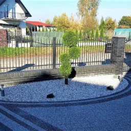 Uformowany krzew w kształcie trzech kul rosnący na białym żwirze w okrągłym klombie przed czarnym, kutym ogrodzeniem z ceglanymi słupkami, widok na dom z balkonem w tle.