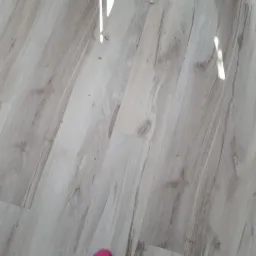 Jasna podłoga panelowa imitująca drewno, ułożona wzdłuż, z widocznymi fugami i refleksami światła, fragment różowej stopy w rogu kadru.