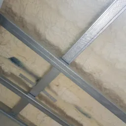 Konstrukcja sufitu podwieszanego z profilami metalowymi wypełniona jasną pianką izolacyjną o nierównej powierzchni, widoczne ślady niebieskiej farby.