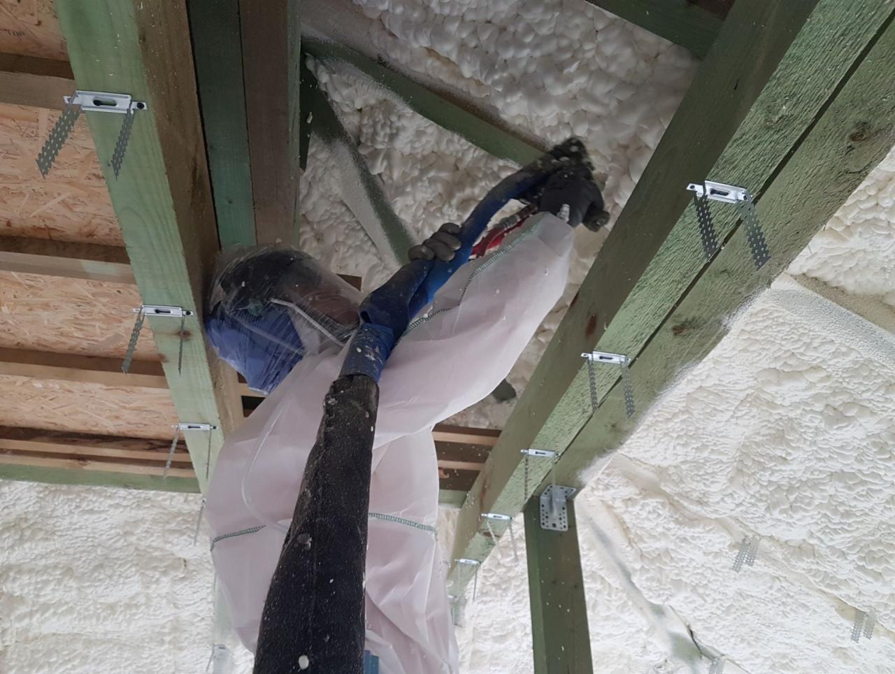 Pracownik w kombinezonie ochronnym aplikuje pianę izolacyjną na poddaszu, widoczne drewniane belki konstrukcyjne z metalowymi łącznikami.