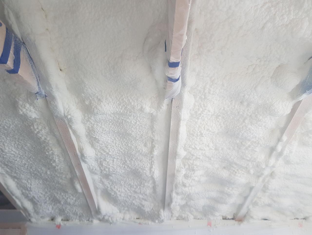 Poddasze ocieplone białą pianką natryskową, widoczne drewniane belki konstrukcyjne i fragmenty zabezpieczonych rur instalacyjnych.