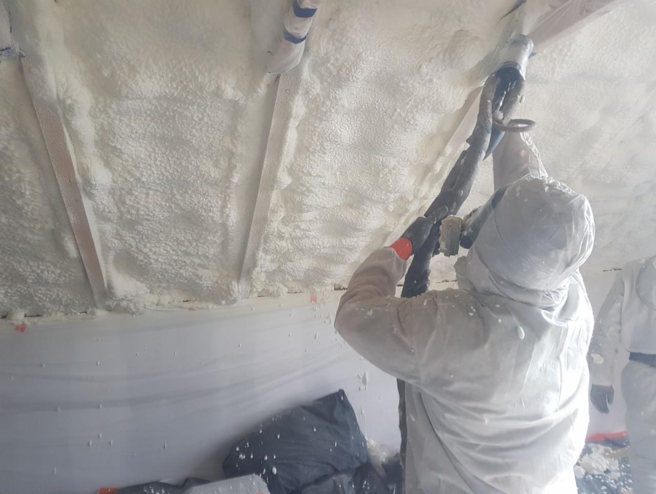 Pracownik w kombinezonie ochronnym aplikuje pianę izolacyjną na strop budynku, widoczne elementy konstrukcyjne i instalacje.