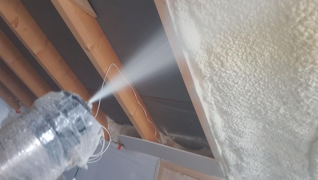 Aplikacja pianki PUR na poddaszu, widoczne drewniane belki stropowe, świeżo nałożona warstwa izolacji o nierównej strukturze i dysza aplikująca pianę.