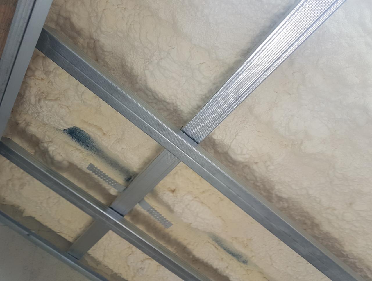 Konstrukcja sufitu podwieszanego z profilami metalowymi wypełniona jasną pianką izolacyjną o nierównej powierzchni, widoczne ślady niebieskiej farby.