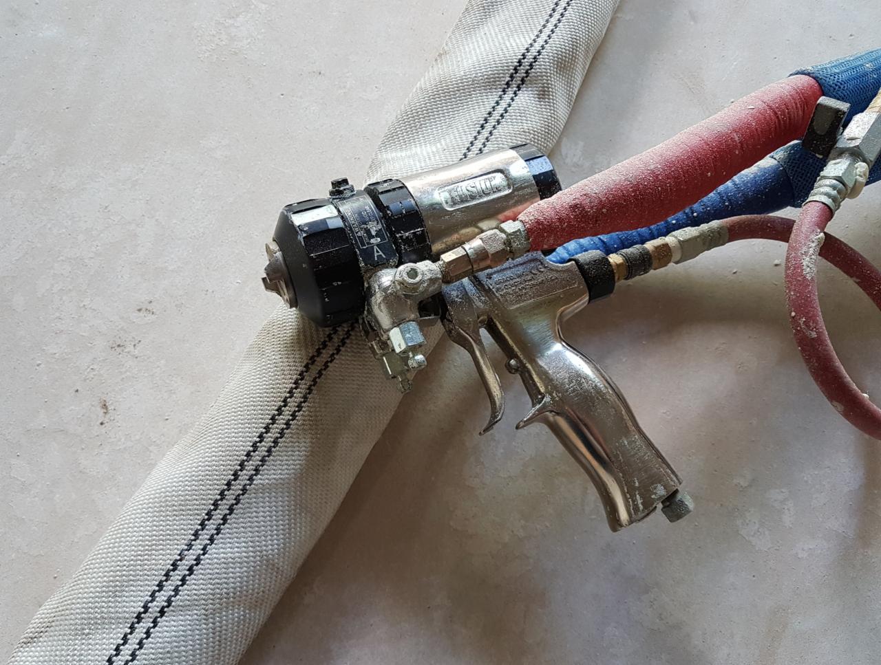 Metalowy pistolet natryskowy z przewodami, używany do aplikacji izolacji, leży na szarym podłożu obok grubego, materiałowego węża. Widoczne ślady użytkowania i zabrudzenia na sprzęcie.