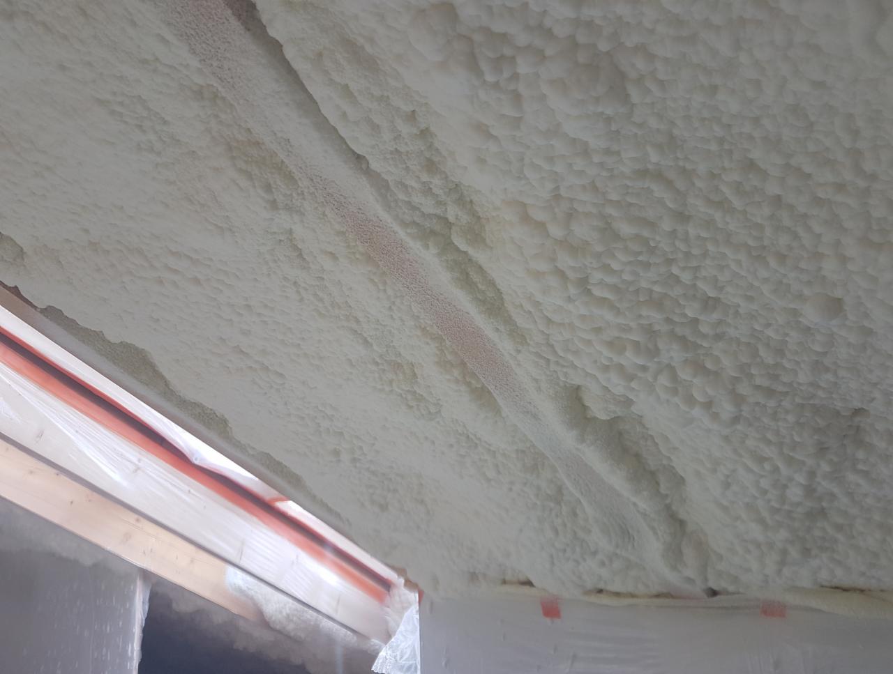 Nierównomierna warstwa białej pianki PUR naniesiona na sufit, widoczne drewniane elementy konstrukcyjne zabezpieczone folią.