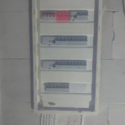 Rozdzielnica elektryczna z zabezpieczeniami różnicowoprądowymi i nadprądowymi zamontowana na surowej ścianie z pustaków, z widocznymi przewodami w białych rurkach PCV u góry i u dołu.