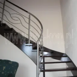 Ciemne, drewniane schody z zakręconą, nowoczesną balustradą ze stali nierdzewnej, widoczne z dołu, z fragmentem zielonej sofy w rogu.