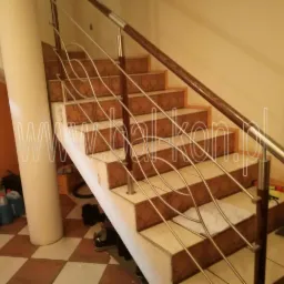 Schody z brązowymi płytkami, balustrada z drewnianą poręczą i wypełnieniem z nierdzewnych prętów o falistym kształcie, widoczne narzędzia montażowe.