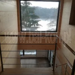 Wnętrze drewnianego domu z widokiem na jezioro przez okno, schody z metalową barierką z poziomymi prętami, nałożony znak wodny na zdjęciu.