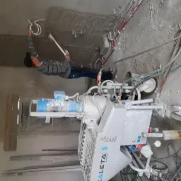 Pracownik w czapce nanosi tynk maszynowy na sufit za pomocą pistoletu natryskowego, obok stoi maszyna do tynkowania Kaleta 5.