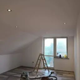 Pokój na poddaszu po malowaniu ścian na biało, widoczne ślady po remoncie: drabina, odkurzacz przemysłowy, wiadro i butelki po farbach. W suficie zamontowane halogeny.