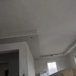 Wnętrze w trakcie prac wykończeniowych; widoczny sufit z zamontowaną konstrukcją podwieszaną z płyt gipsowo-kartonowych i nałożoną warstwą gładzi, częściowo zakryty sylwetką osoby w roboczym ubraniu.