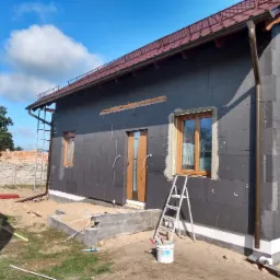 Dom jednorodzinny w trakcie ocieplania styropianem grafitowym, widoczne okna z drewnianą stolarką, brązowe rynny oraz schody wejściowe w surowym betonie i drabina oparta o ścianę.